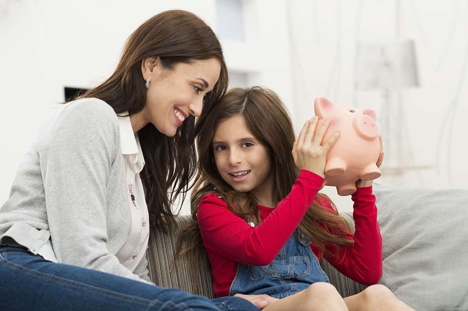 Smiling Mother Looking At Her Daughter Sitting On Couch Holding Piggybank